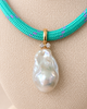 A close-up of a turquoise woven necklace with small purple accents and a large baroque pearl pendant set in gold, displayed on a beige fabric jewelry bust.