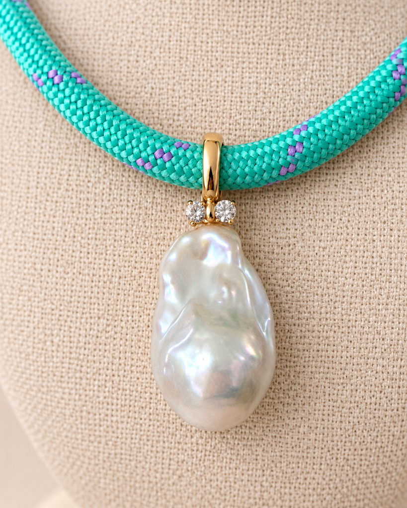 A close-up of a turquoise woven necklace with small purple accents and a large baroque pearl pendant set in gold, displayed on a beige fabric jewelry bust.