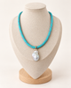 A turquoise woven necklace with a large white baroque pearl pendant, displayed on a beige fabric jewelry bust with a wooden base against a light cream background.