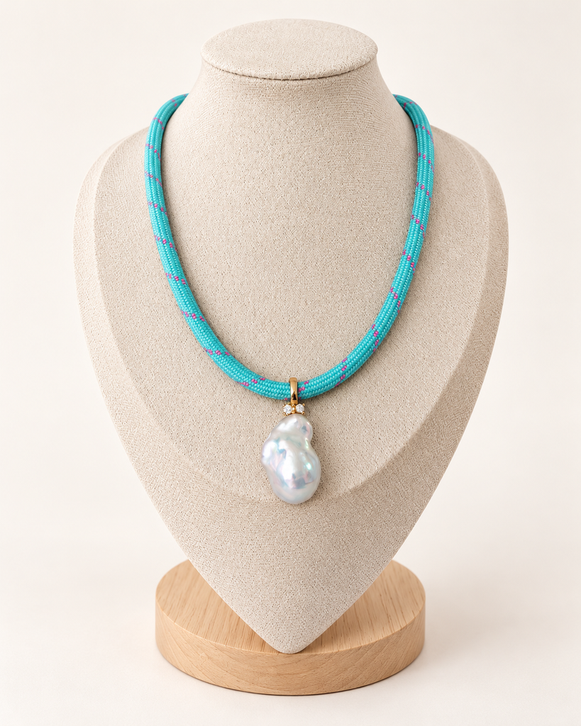 A turquoise woven necklace with a large white baroque pearl pendant, displayed on a beige fabric jewelry bust with a wooden base against a light cream background.