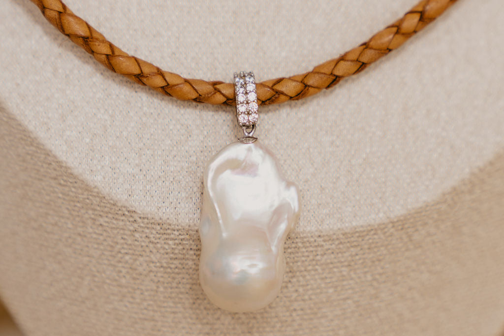 Pearl pendant with diamond accents on a braided leather cord against a beige fabric background
