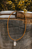 Brown braided leather necklace with a freshwater pendant on a rustic metal surface with yellow flowers in the background