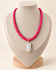 A bright pink braided cord necklace with a white baroque pearl pendant and a small gold accent, displayed on a beige fabric jewelry bust.