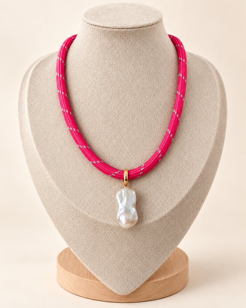 A bright pink braided cord necklace with a white baroque pearl pendant and a small gold accent, displayed on a beige fabric jewelry bust.