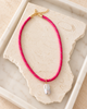 A bright pink cord necklace with small gold speckles and a gold clasp, featuring a single irregular white pearl pendant, displayed on a textured stone slab inside a white tray.