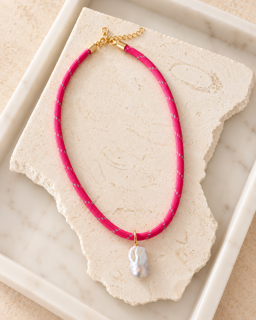 A bright pink cord necklace with small gold speckles and a gold clasp, featuring a single irregular white pearl pendant, displayed on a textured stone slab inside a white tray.