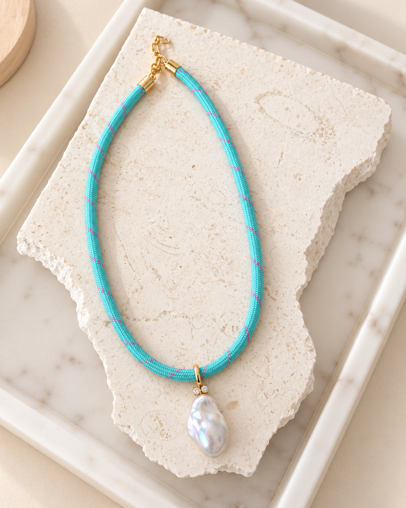 A turquoise woven necklace with a large white baroque pearl pendant, displayed on a beige fabric jewelry bust with a wooden base against a light cream background.