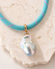 A turquoise woven necklace with a large white baroque pearl pendant, displayed on a beige fabric jewelry bust with a wooden base against a light cream background.